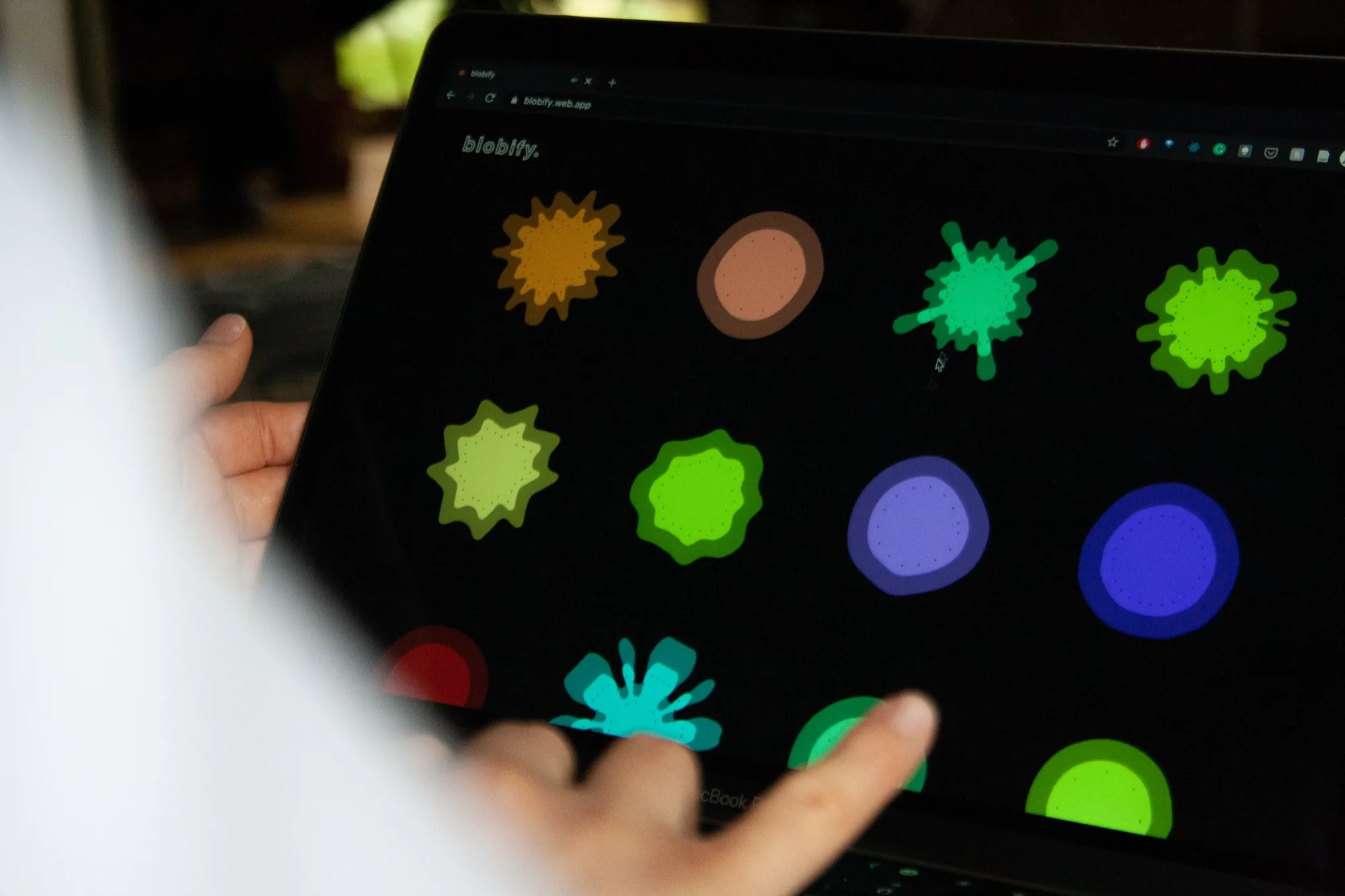 A laptop screen filled with coloured blobs of different shapes, a hand reaching toward one