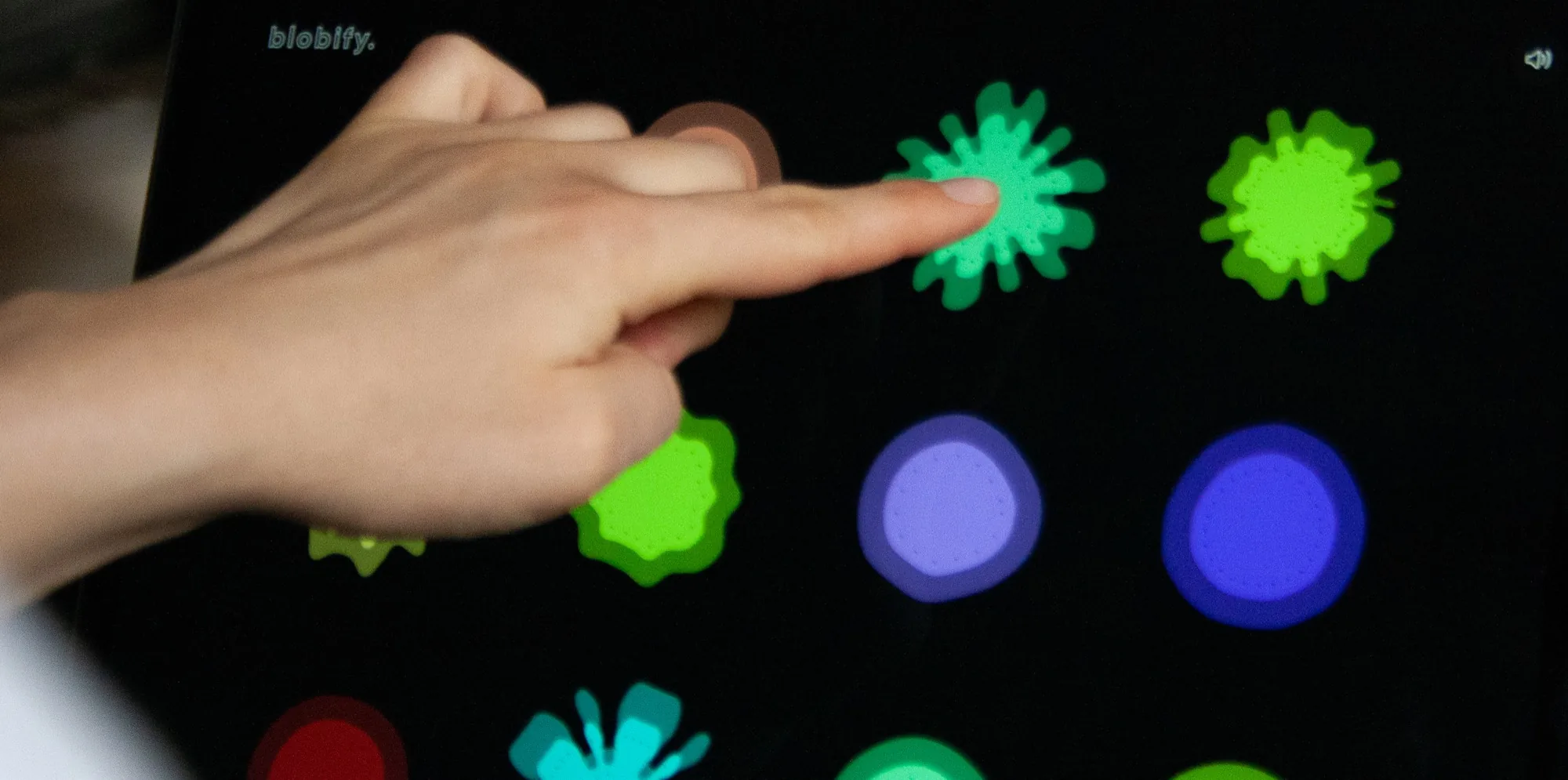 A finger touching a spiky green blob on a dark screen, surrounded by other coloured blobs