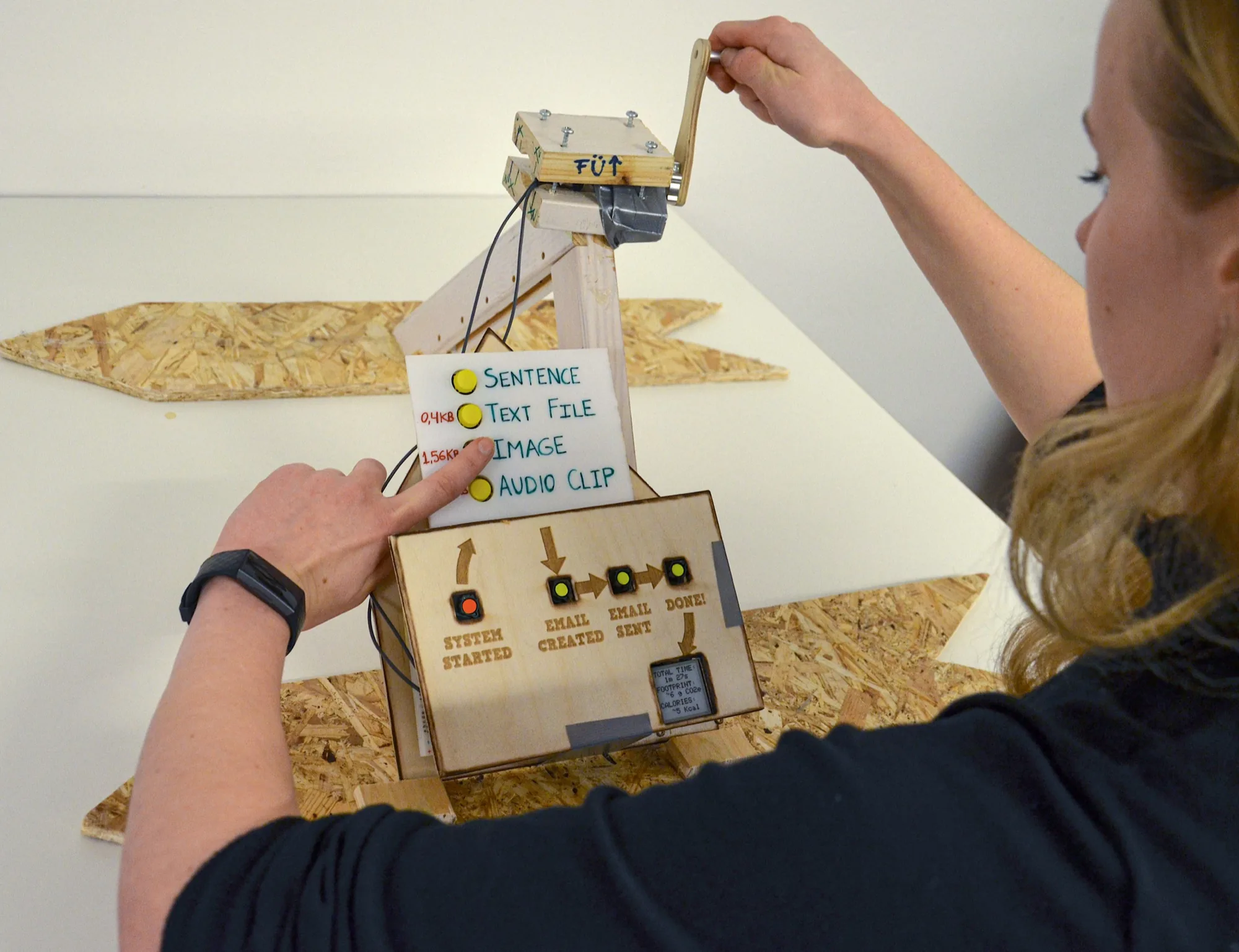 A person turning a wooden hand crank on the email machine while selecting a message type on its panel