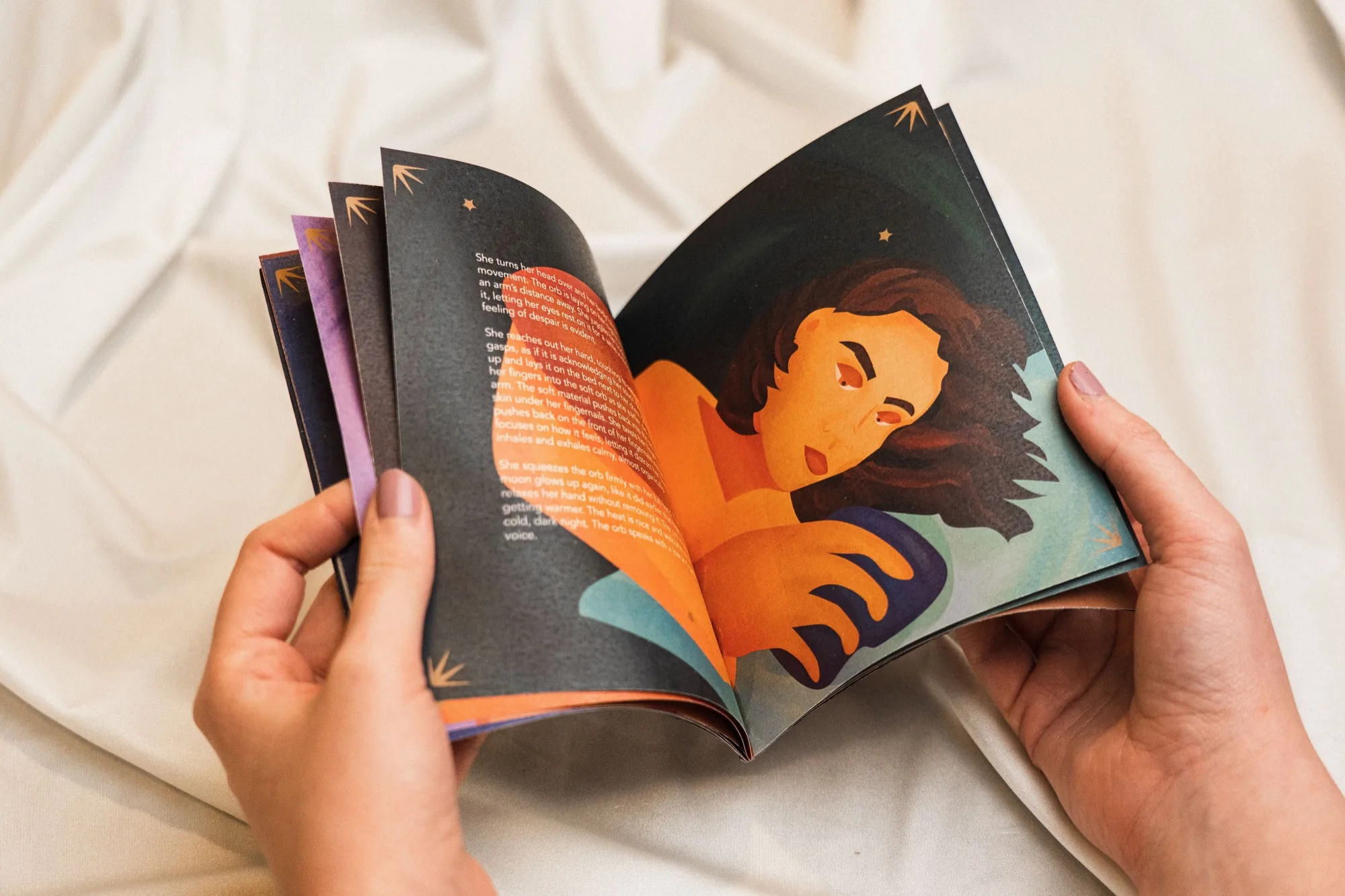 Hands holding open the illustrated booklet, showing a woman lying awake at night in orange and teal